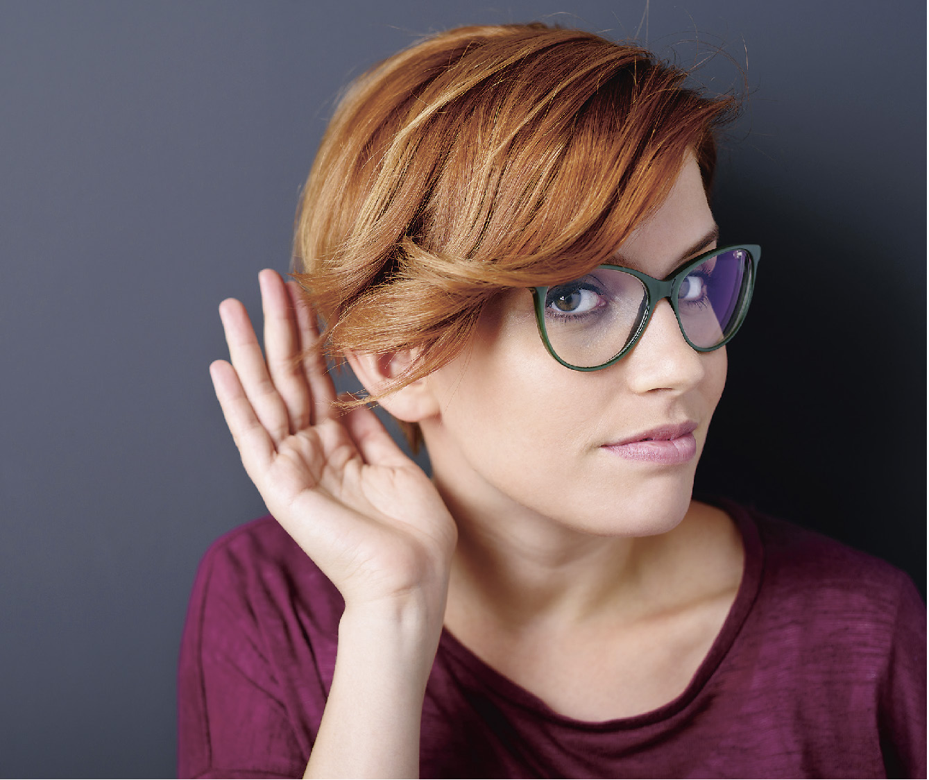 Frau mit roten Haaren und Brille hält sich Hand an das Ohr